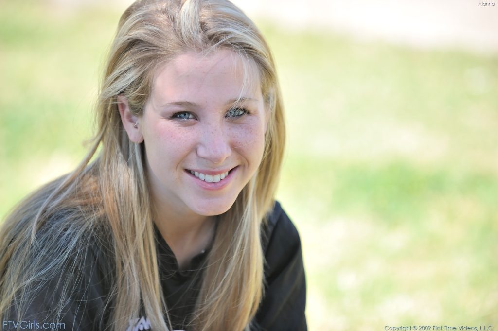 A young woman with long, blonde hair smiles at the camera while seated outdoors. The background features green grass, adding a natural touch to the scene.